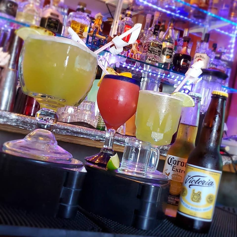 Margaritas and Mexican beers on a bar with bottles of liquor and purple lighting in the background.