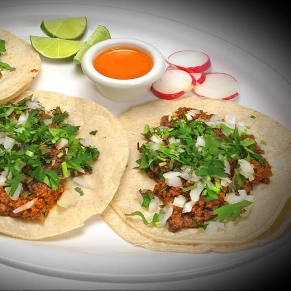 Three La Casita Mexican tacos with meat, cilantro, and onions, served with lime, radishes, and sauce.